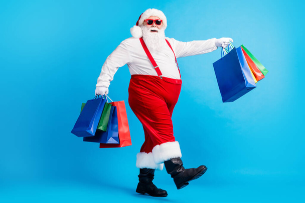 Full length profile side photo of crazy modern white grey hair bearded santa claus go shopping buy x-mas christmas bags wear red suspenders overalls boots isolated blue color background - Fotó, kép