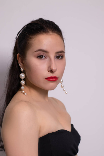 Emotionales Porträt einer attraktiven Brünetten in schwarzem Top mit weißen Perlenohrringen auf weißem Hintergrund - Foto, Bild