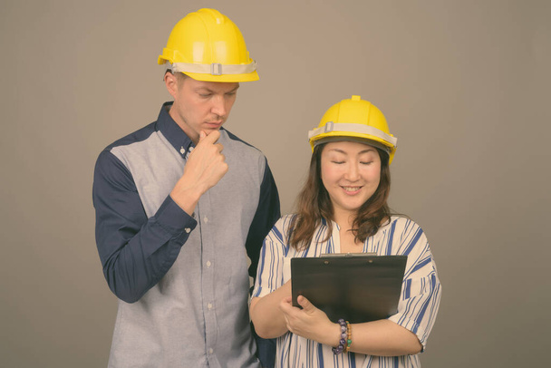 Young handsome businessman and mature Asian businesswoman wearing hardhat against gray background - Photo, Image