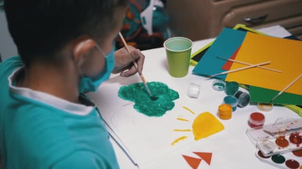 Kid in Mask Senta-se à Mesa no Quarto e Tintas Imagem com Pincel com Pintura Verde - Filmagem, Vídeo