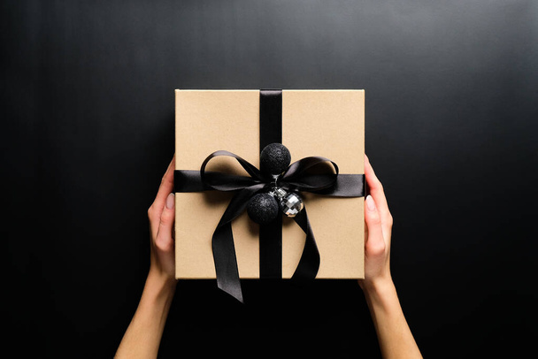 Female hands holding cardboard gift box wrapped black ribbon and Christmas balls over black background. Christmas present, New Year gift box concept. - Photo, Image
