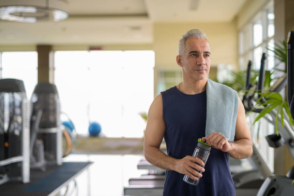 Porträt eines reifen, gut aussehenden persischen Mannes mit Wasserflasche im Fitnessstudio - Foto, Bild