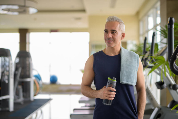 Maduro guapo persa hombre pensando mientras sostiene la botella de agua en el gimnasio - Foto, imagen