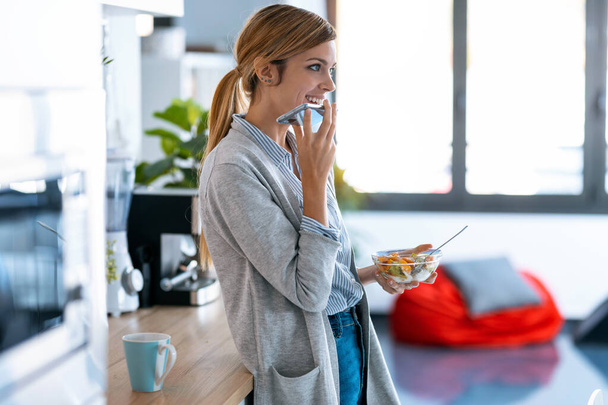 Shot of beautiful young woman sending audio with mobile phone while having healthy breakfast in the kitchen at home.  - Foto, Imagem