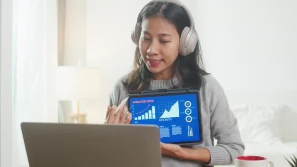 Ázsiai üzletasszony visel fejhallgató segítségével laptop beszélni kollégák tervről videohívás, míg okos dolgozik otthonról a hálószobában. Önizolálás, társadalmi távolság, karantén a koronavírus számára. - Felvétel, videó
