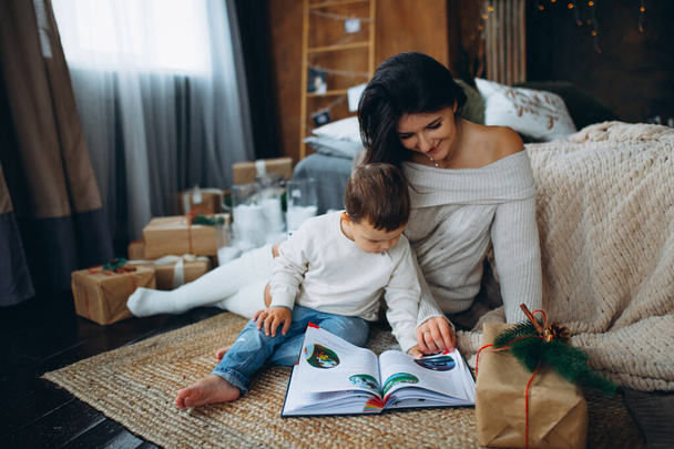 beautiful brown-haired mom and lovely boy son sitting on the floor and reading a book of fairy tales on different christmas tree. - Photo, image