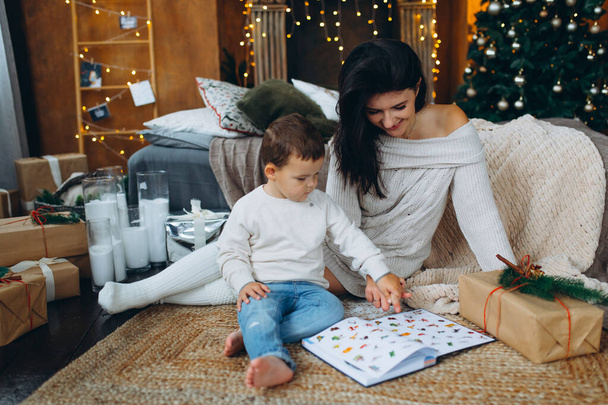 beautiful brown-haired mom and lovely boy son sitting on the floor and reading a book of fairy tales on different christmas tree. - Foto, afbeelding