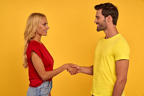 Side view of smiling young couple friends guy girl in colored t-shirts isolated on yellow wall background studio portrait. People lifestyle concept. Mock up copy space. Holding greeting with hands - Foto, Imagen