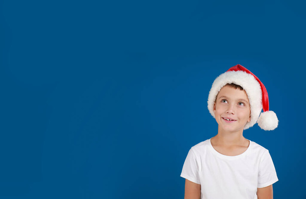 Little child in Santa hat dreaming on blue background, space for text. Christmas celebration - Фото, изображение