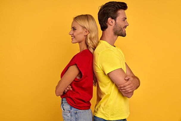 Boční pohled na usmívající se mladý pár v červených a žlutých tričkách izolovaných na žlutém pozadí. Koncept životního stylu lidí. Zesměšnit kopírovací prostor. Stojíme zády k sobě, držíme se za ruce - Fotografie, Obrázek
