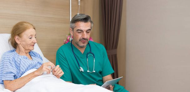 Lekarz rehabilitacyjny trzymający tabletkę cyfrową opisuje możliwości leczenia kobiet w podeszłym wieku cierpiących na osłabienie mięśni, trudności w chodzeniu podczas leżenia w szpitalnym łóżku.. - Zdjęcie, obraz