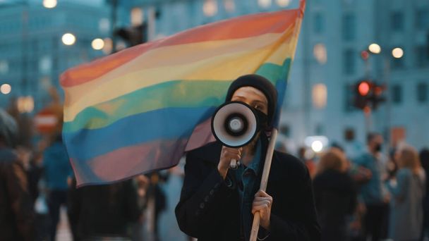 Junge Frau mit Gesichtsmaske spricht ins Megafon und hält Regenbogenfahne in der Menge - Foto, Bild