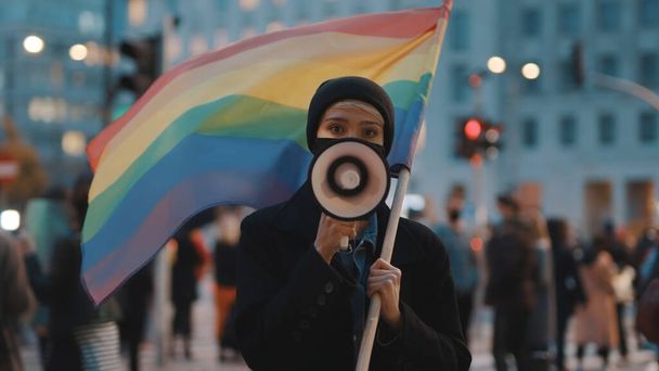 Молодая женщина в маске говорит в мегафон, держа в толпе радужный флаг - Фото, изображение