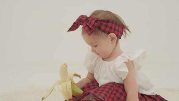 Egy kislány kockás fejpántban és szoknyában nagy hámozott banánt tart. Lassított felvétel lezárva. - Felvétel, videó