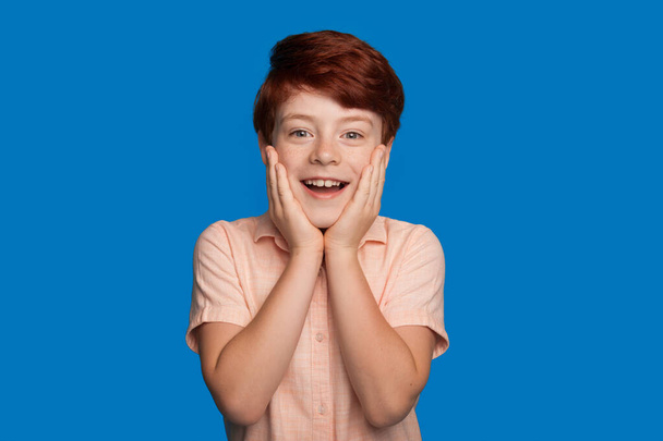 Σοκαρισμένο καυκάσιο αγόρι αγγίζει το πρόσωπό του ενώ κοιτάζει την κάμερα και χαμογελά σε ένα μπλε τοίχο στούντιο - Φωτογραφία, εικόνα