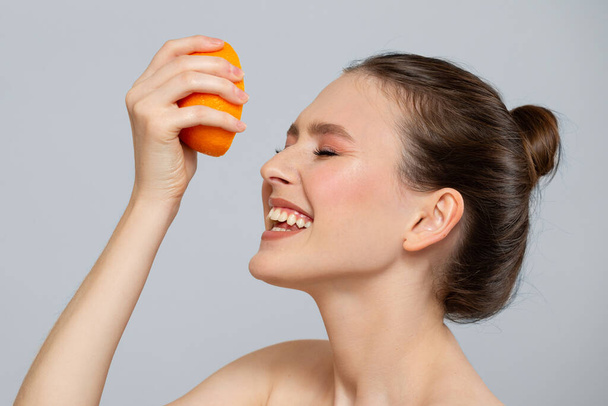 Śmiejąca się kobieta z pięknym uśmiechem i czystą skórą przynosi jej pół pomarańczy na twarz. Leczenie owoców witaminami na twarz - Zdjęcie, obraz