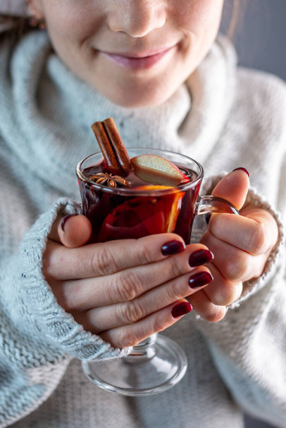 A woman in a white warm sweater is holding a transparent glass cup of mulled wine in her hand. Concept of a cozy atmosphere and a traditional winter drink. - Valokuva, kuva