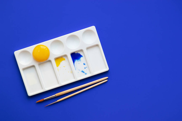 White palette with watercolor paints and brushes. Blue background. Instead of yellow, the egg yolk is used. Creative design and art. Top view and copy space - Photo, Image