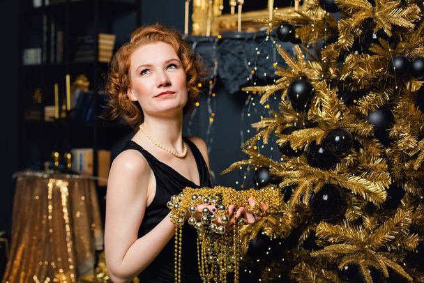 Genç bir kadın, elinde altın süslemelerle bir Noel ağacının arka planında boncuk dizisi tutuyor. Şık kadınlar, şık gece kıyafetleri, aksesuarlar, makyaj. Mutlu yıllar.. - Fotoğraf, Görsel
