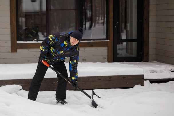 Человек с лопатой очищает двор своего деревянного серого дома от снега. Селективный фокус. - Фото, изображение
