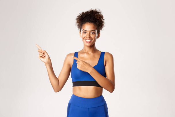 Femme de fitness afro-américaine attrayante et en bonne santé, portant une tenue de sport bleue, pointant du doigt le coin supérieur gauche et souriant, montrant le logo, fond blanc - Photo, image