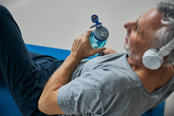 Hombre mayor positivo con una botella de agua escuchando música - Foto, imagen