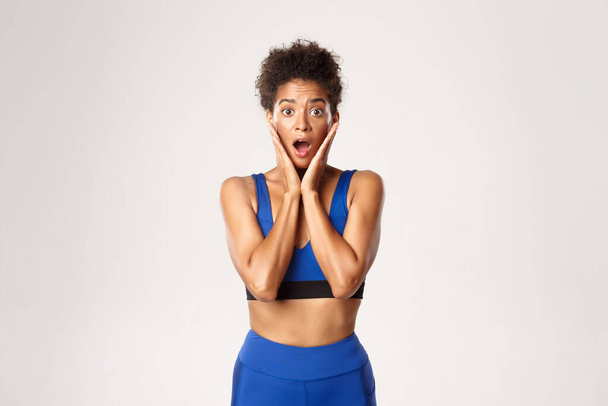 Cintura-up de surpreso afro-americano ajuste feminino athelte, olhando espantado, boca aberta maravilhada, de pé contra fundo branco em azul esporte roupa - Foto, Imagem