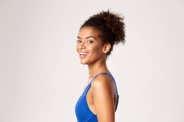Concept of workout and sport. Close-up profile shot of happy, athletic woman in blue fitness clothing, turn head at camera and laughing, standing against white background - Photo, Image