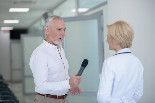 Gray-haired male journalist interviewing blonde female doctor in corridor - Foto, Imagen