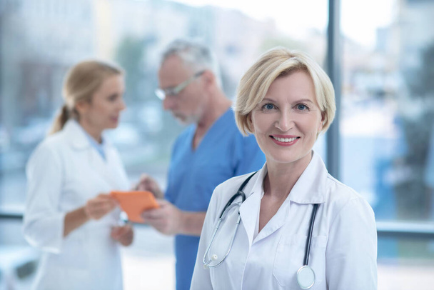 Mature female doctor smiling into camera, her colleagues standing with tablet behind - Foto, Imagem