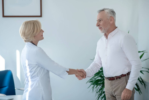Gray-haired male patient shaking hands with blonde female doctor - Фото, изображение