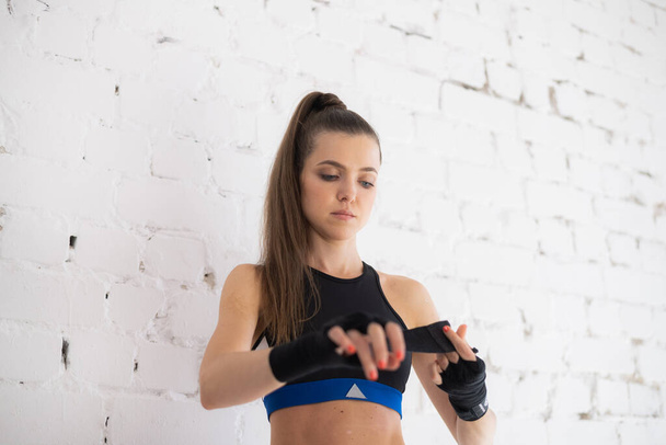 Egy női bokszoló egy fehér téglafalhoz teker egy sportkötést, amiért bokszol a kezén. - Fotó, kép