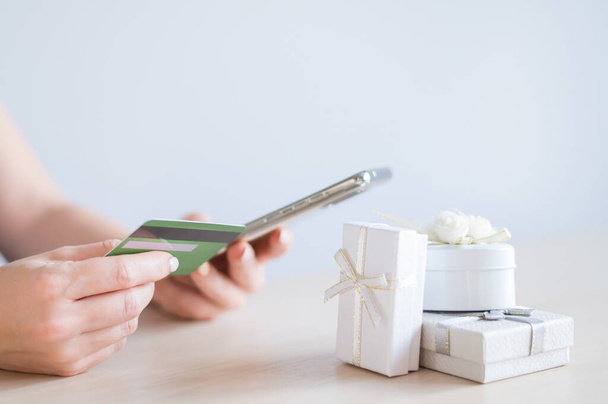 A faceless woman sits at a table and holds a mobile and a credit card in her hands. Online shopping for gifts concept - Φωτογραφία, εικόνα