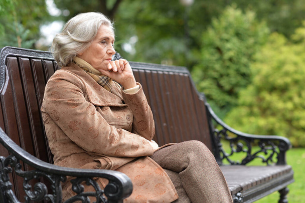грустная, заботливая пожилая женщина, сидящая на скамейке в парке - Фото, изображение
