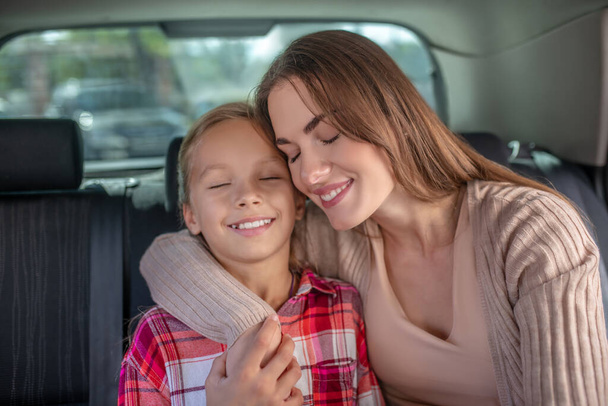 Mom embracing her daughter, sitting cheek to cheek on backseat of car - Foto, imagen