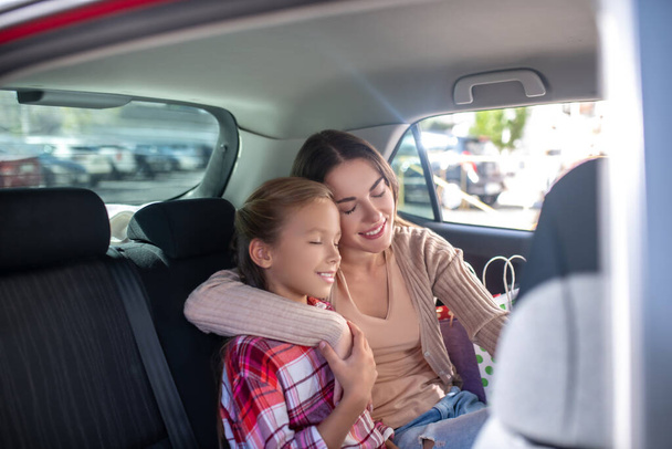 Mom hugging her daughter, sitting cheek to cheek on backseat of car - Photo, image