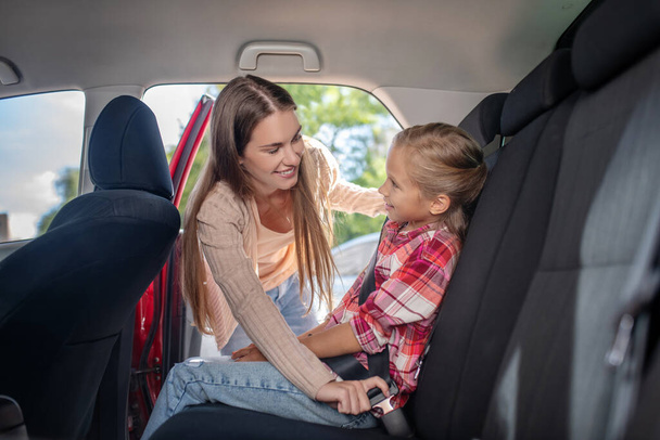 Smiling mom fastening her daughters safety belt on backseat of car - Фото, изображение