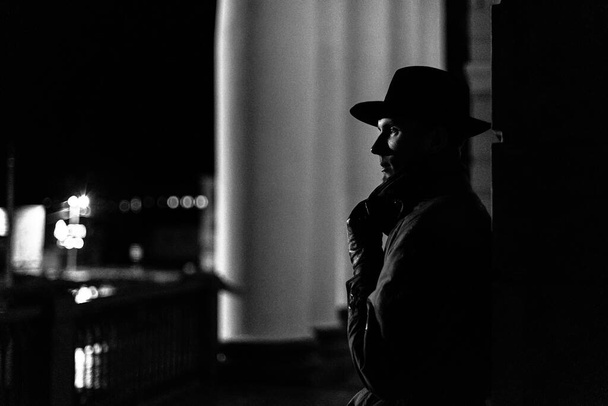 donker silhouet van een man in een regenjas met een hoed en een litteken op zijn gezicht 's nachts - Foto, afbeelding