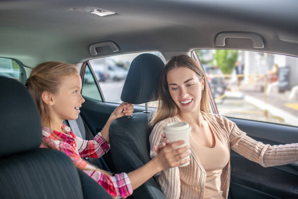 Girl giving coffee cup to her mom sitting at the wheel - Photo, image