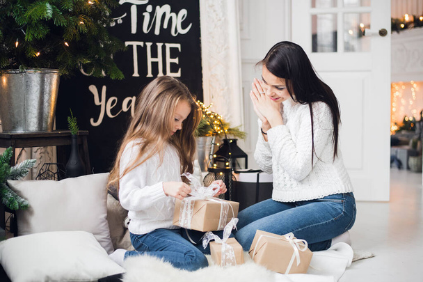 Merry Christmas and Happy Holidays. Cheerful mom and her cute daughter girl opening a Christmas present. Parent and little child having fun near Christmas tree indoors - Fotó, kép