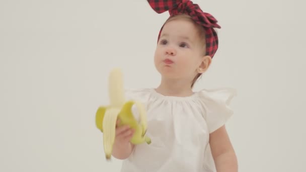 再生ヘッドバンドやスカートの中の小さな女の子は大きなバナナを食べている。スローモーションを閉じます - 映像、動画
