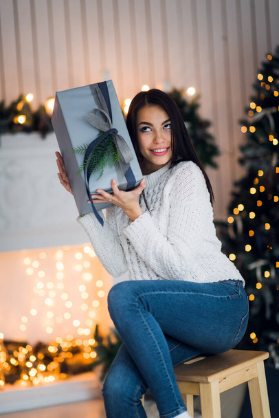 Счастливая молодая женщина с рождественским подарком удивлен и взволнован сидя дома против рождественских украшений - Фото, изображение