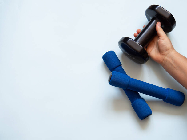Two blue small dumbbells and a caucasian woman hand grabbing one black dumbbell for weight and strength training in white background. Copy space for text. Weightlifting and healthy lifestyle concept - Foto, afbeelding