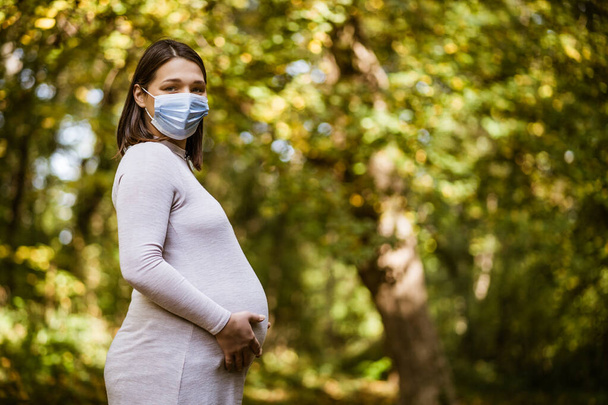 Portrét těhotné ženy v parku na podzim. Má na sobě ochrannou masku. Covid-19 pandemický koncept. - Fotografie, Obrázek