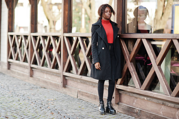 Portrait of a curly haired african woman wearing fashionable black coat and red turtleneck posing outdoor. - Foto, immagini