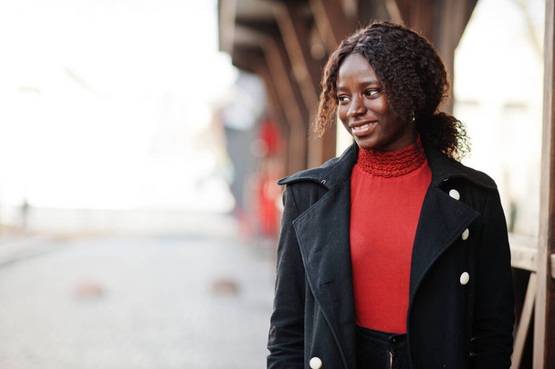 Portrait of a curly haired african woman wearing fashionable black coat and red turtleneck posing outdoor. - Foto, afbeelding