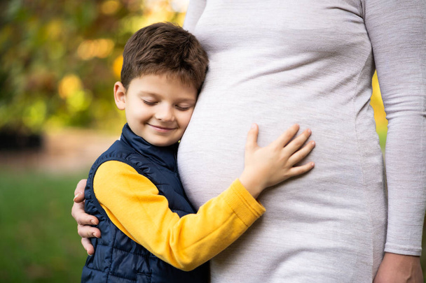 男の子は秋に公園で妊婦の胃にもたれている。自然の中で家族のリラックスした時間. - 写真・画像