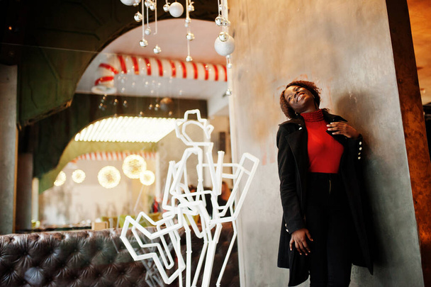 Portrait of a curly haired african woman wearing fashionable black coat and red turtleneck posing at cafe. - Foto, afbeelding