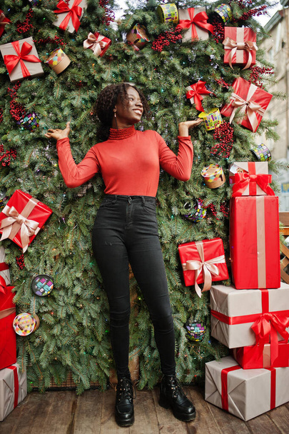 Portrait of a curly haired african woman wearing fashionable red turtleneck posing against christmas decorations, new year eve theme. - Photo, image