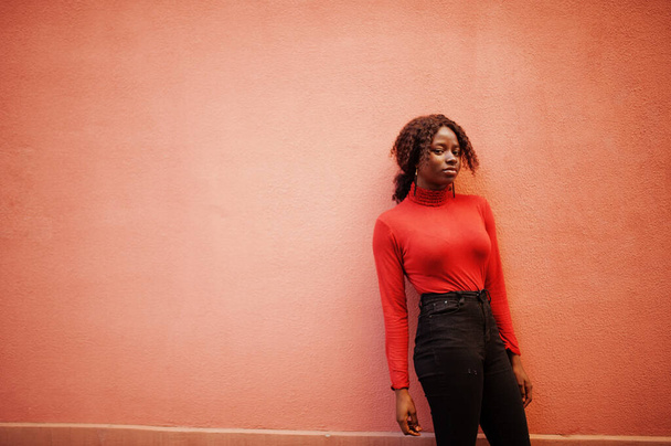 Portrait of a curly haired african woman wearing fashionable red turtleneck posing against rose wall. - Photo, Image